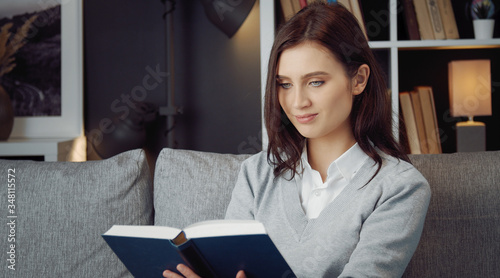 Waist up portrait of young delighted beauty reading book sitting on sofa staying in flat