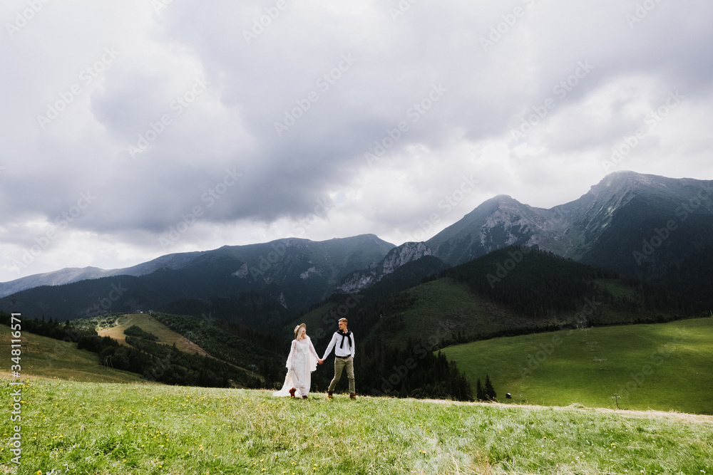 Beautiful bride in a boho style dress and groom walk on the field near the mountains. Wedding photo shoot in the mountains.