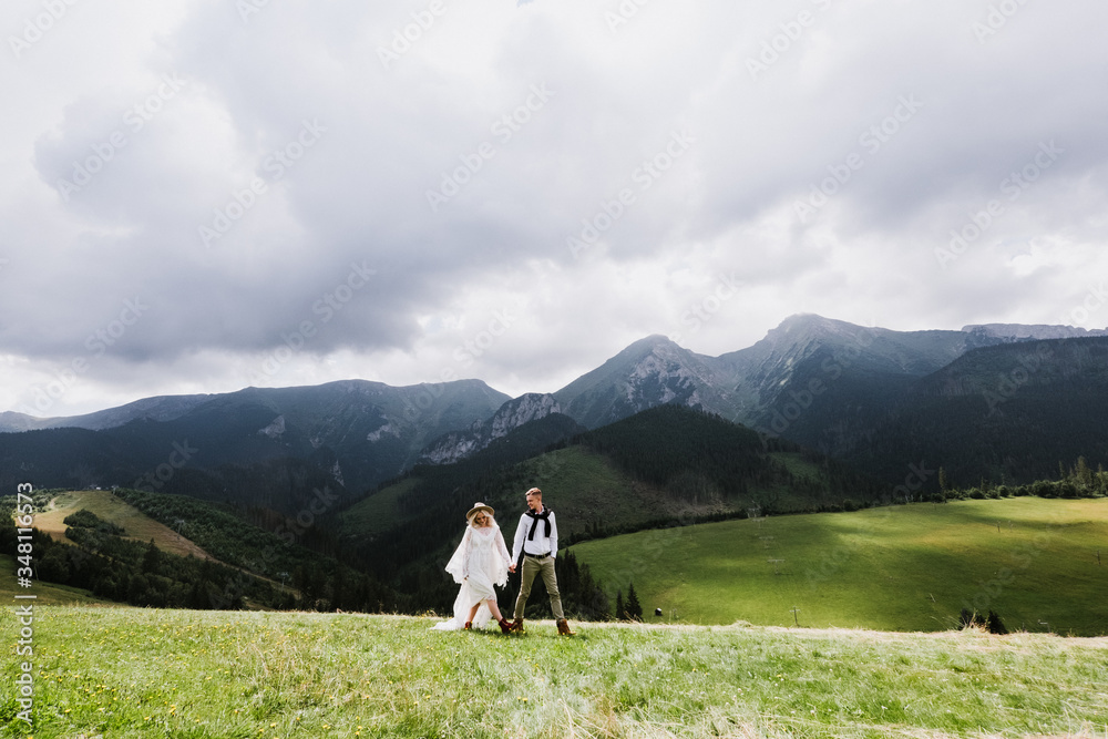 Beautiful bride in a boho style dress and groom walk on the field near the mountains. Wedding photo shoot in the mountains. Beautiful bride in a boho style dress and groom walk on the field near the mountains. Wedding photo shoot in the mountains.