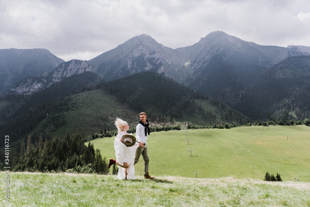 Naklejka premium Beautiful bride in a boho style dress and groom walk on the field near the mountains. Wedding photo shoot in the mountains.