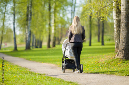 Fototapeta Naklejka Na Ścianę i Meble -  One young mother pushing white baby stroller and slowly walking at town green park in warm, sunny spring day. Spending time with infant and breathing fresh air. Enjoying stroll. Back view.