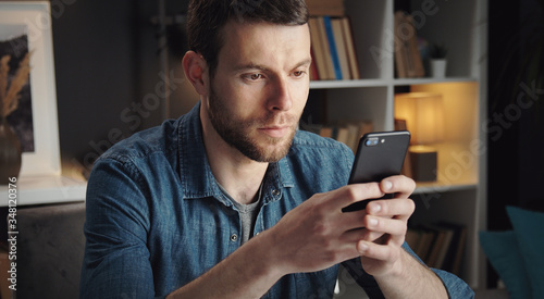 Cropped shot of young man in denim shirt holding smartphone and texting staying in apartment