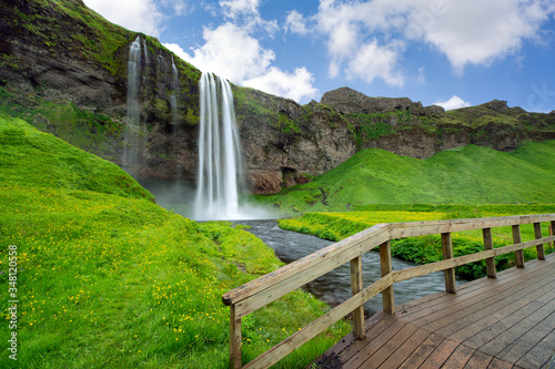 Seljalandfoss falls and streams in the summer, there are green grass and yellow flowers all over the area The front scene has a wooden bridge. This is a popular tourist destination in Iceland.