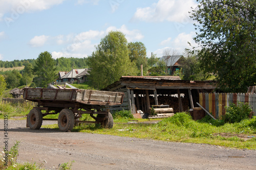 A Russian countryside view with traditional houses on the background. The pile of logs, cart, bushes and trees on the foreground.