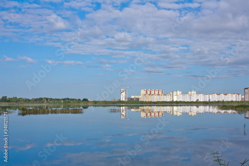 A clean lake with reeds, a blue cloudy sky and the construction of high-rise buildings in the background.