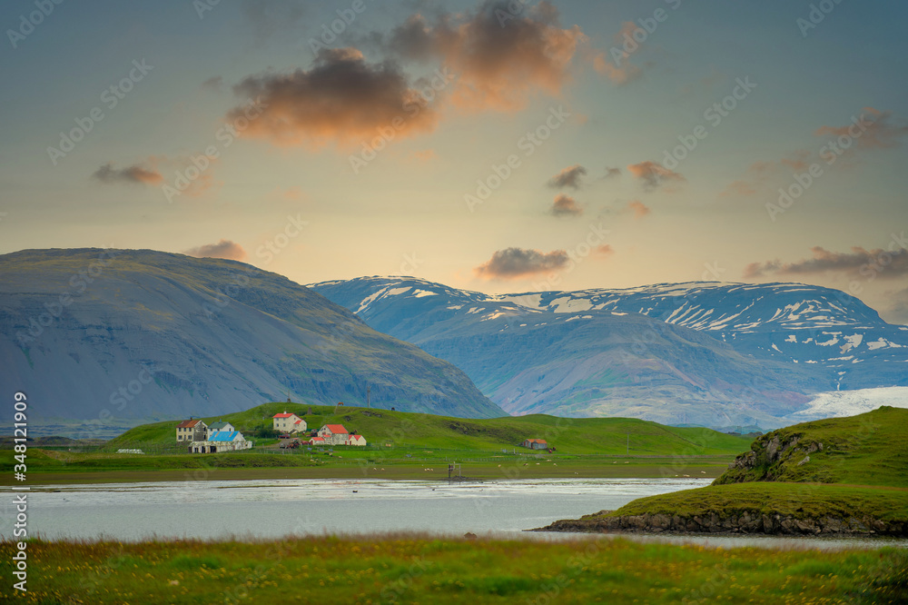 Fototapeta premium The house and ranch are located on a green meadow next to the lake. The background is high mountains, the sunset and beautiful sky, In the countryside in Iceland.