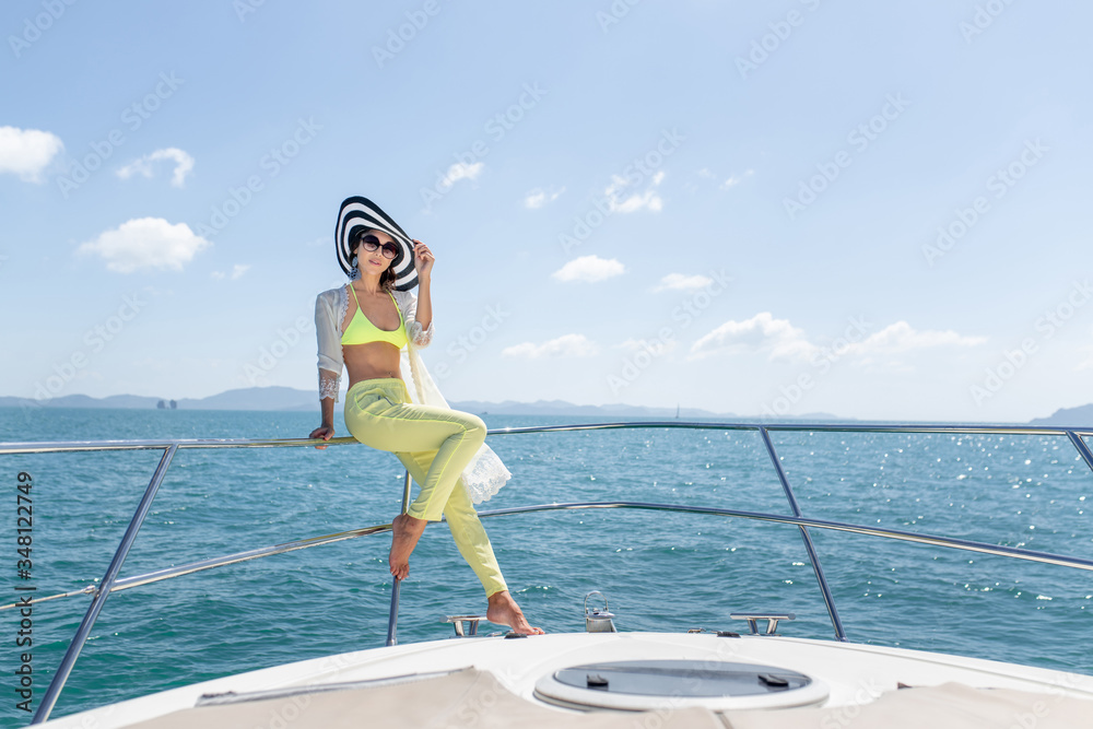 Naklejka premium Outdoor shot of adorable young woman in a summer suit and hat sitting on edge of yacht, looking to beautiful nature landscape during trip. Happy woman enjoying summer travel. Back view