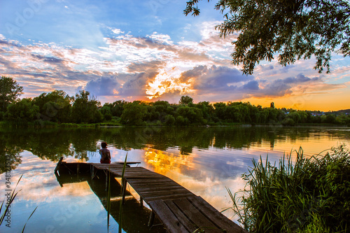 Ukraine. Sunset over the river. There is a lonely figure on the pier.
  The rays of the sun make their way through clouds. Reflected in the river
clouds with the setting sun and trees.