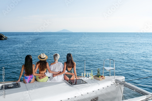 A group of four young women embracing each other are looking at the sea horizon while sitting on the edge of the yacht. Summer holidays. Cruise © Detkov D
