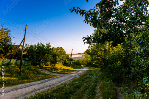 Ukraine. Early morning in the village. Sunrise.
A dirt road runs through the village. A power line has been drawn along the road. The rays of the sun make their way through
  foliage of trees.