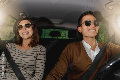 Happy caucasian couple sitting inside car driving, wearing seat belt and sunglasses. Smiling during the travel. © Pintau Studio