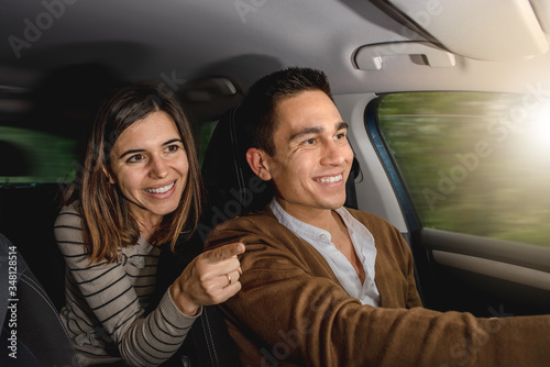 Caucasian couple inside car smiling. Woman is pointing something outside with finger while man is driving. Happy faces in a journey or travel. Moving car with blurred background.