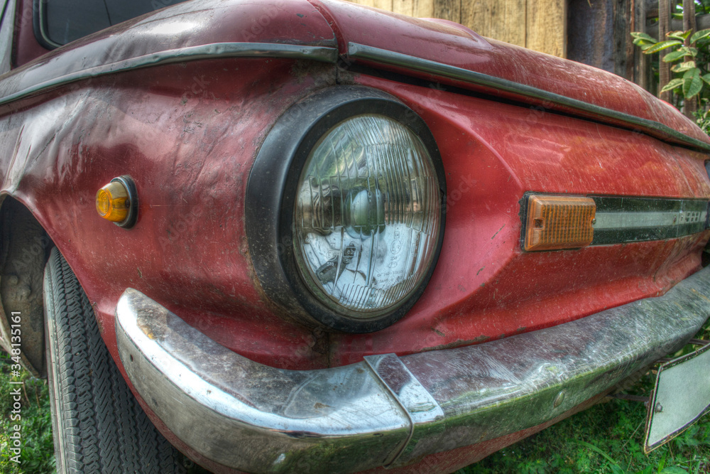 White headlight in a black frame of a red car with a silver bumper and a black wheel, reflections stand on the green grass
