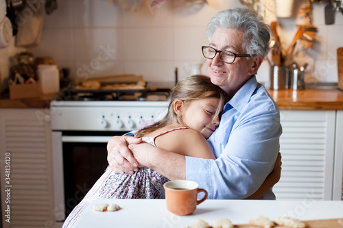 Senior woman hugging child at home. Happy family enjoying kindness, support, care together in cozy kitchen. Cute girl visiting grandmother. Lifestyle moments. Holiday Thanksgiving. photo