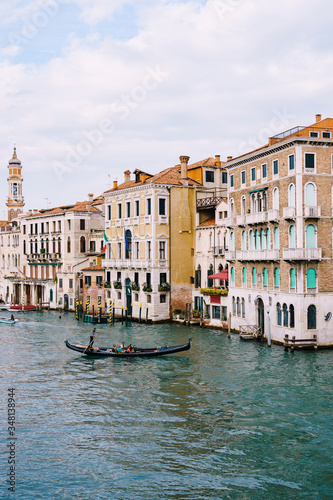 A gondola with tourists is sailing along the Grand Canal amid the facades of Venetian houses standing on the water in Venice, Italy. © Nadtochiy