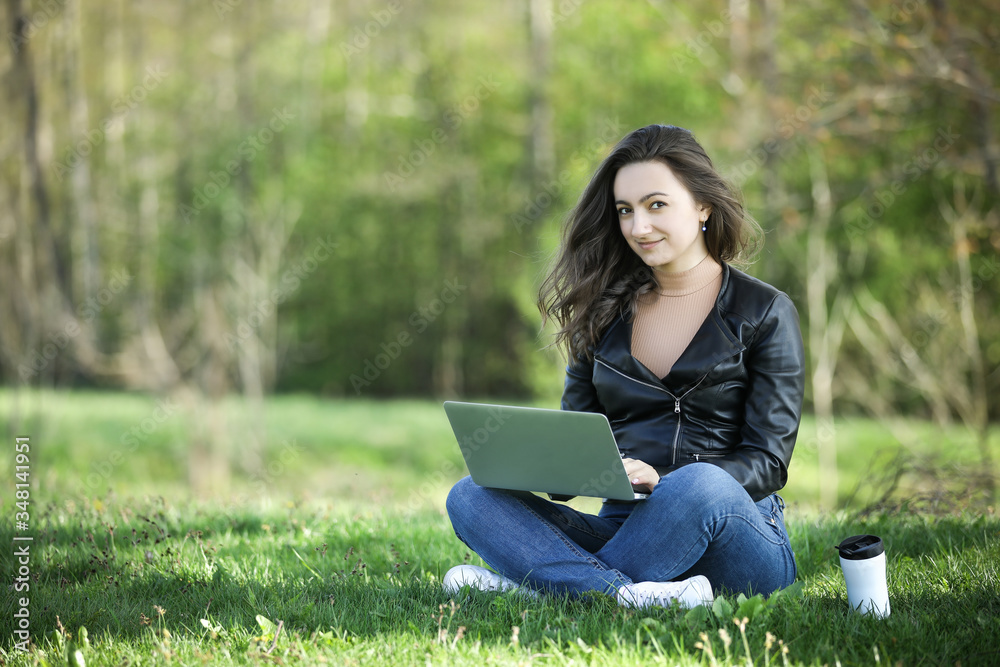 Young woman in a black jacked with coffee cup and laptop working outside in a park. Remote work. Digital work. Distance learning Young woman in a black jacked with coffee cup and laptop working outside in a park. Remote work. Digital work. Distance learning