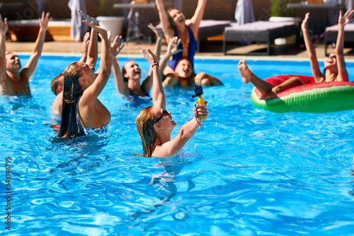 Multiracial group of friends having party in private villa swimming pool. Happy young people in swimwear dancing and splashing with inflatable floaties at luxury resort on sunny day. Girls in bikini.