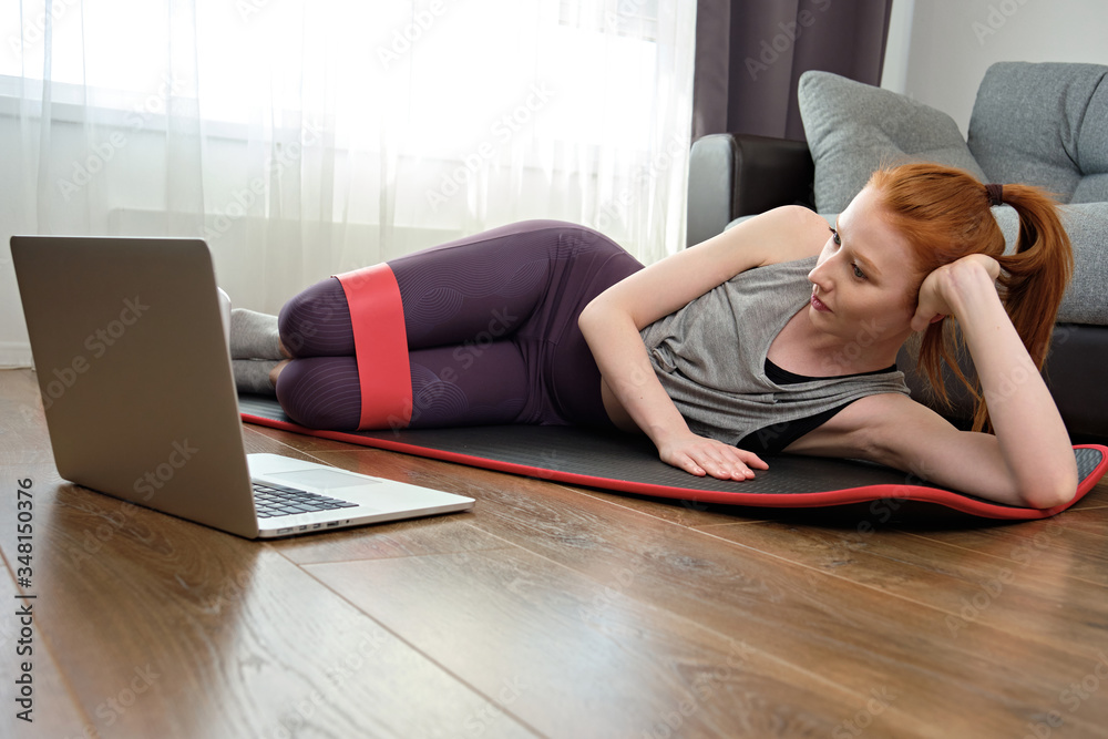 The red-haired girl lies on the sports mat on her side, with an elastic band on her hips, and looks at her laptop. The red-haired girl lies on the sports mat on her side, with an elastic band on her hips, and looks at her laptop.