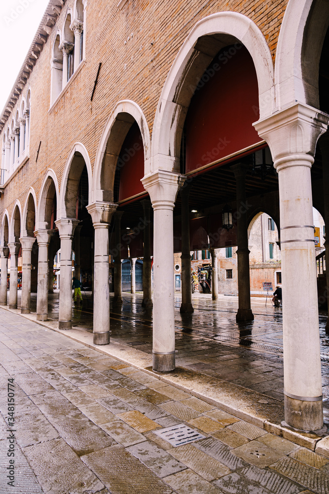 A building in Venice standing on columns. Stone wall of brown brick, white columns and high arches, ancient paving tile.