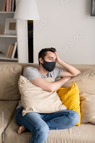 Serious young man in depression wearing cloth mask leaning head on hand while sitting on sofa during quarantine © pressmaster