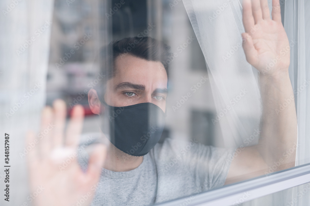 Behind glass view of sad young man in black protective mask holding hands on window and contemplating outdoor scape, he wants to get out of house Behind glass view of sad young man in black protective mask holding hands on window and contemplating outdoor scape, he wants to get out of house