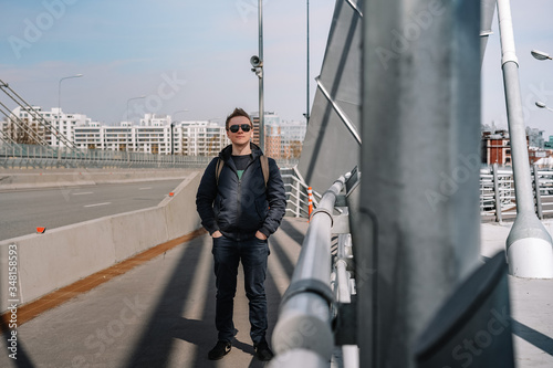 A young man in a jacket and glasses stands on a bridge, beautiful metal structures in the air, hair flying in the wind, business issues, construction engineering photo