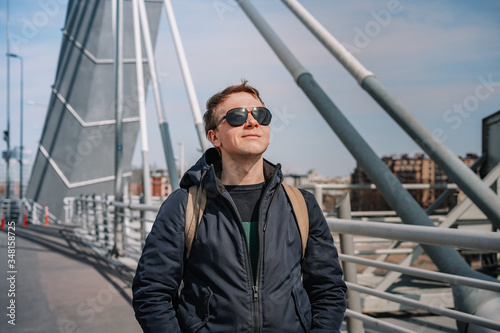 A young man in a jacket and glasses stands on a bridge, beautiful metal structures in the air, hair flying in the wind, business issues, construction engineering