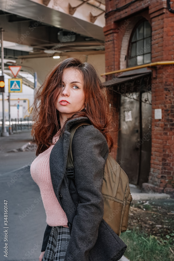 Portrait of a girl with long hair in a coat and a backpack standing on a street in St. Petersburg, a brick old building and bridge beams located close to a residential building