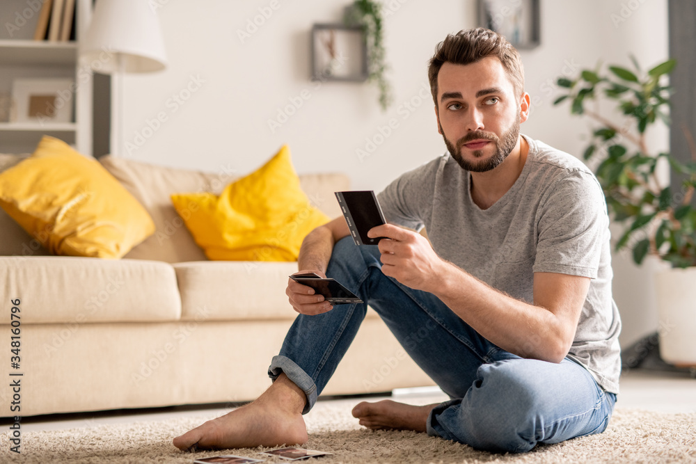 Serious young bearded man sitting on floor in living room and watching photos while spending time at home in quarantine