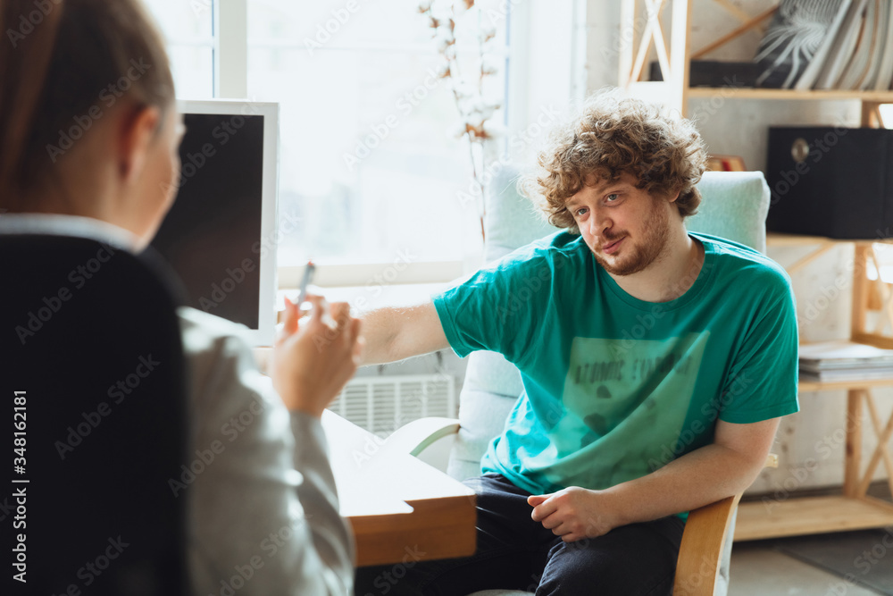 Obraz premium Young man sitting in office during the job interview with female employee, boss or HR-manager, talking, thinking, looks confident. Concept of work, getting job, business, finance, communication.