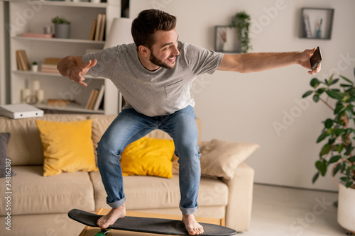 Positive young bearded man balancing on skateboard on coffee table and shooting video for social media at home