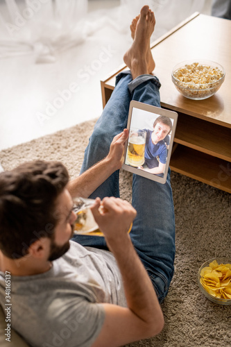 Young man sitting on floor and using tablet while toasting with friend via telecommunication app photo