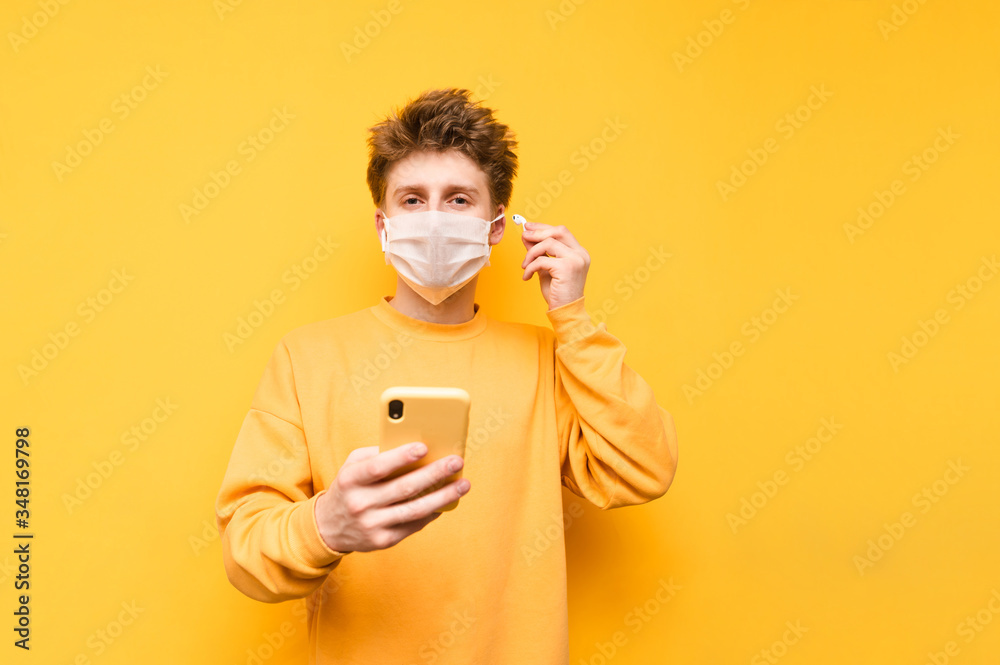 A young man in a protective mask stands on a yellow background with a smartphone in his hand, listens to music in wireless headphones and looks at the camera. Coronavirus pandemic. COVID-19 A young man in a protective mask stands on a yellow background with a smartphone in his hand, listens to music in wireless headphones and looks at the camera. Coronavirus pandemic. COVID-19