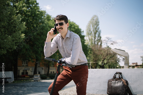 Happy man wearing sunglasses speaking on mobile phone. Smiling man with facial hair talking on the phone while standing outdoor with scooter.