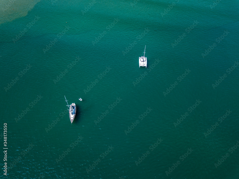 A view from a high-angle drone sees sailboats leaving anchors in a beautiful bay, clear glass-like water.