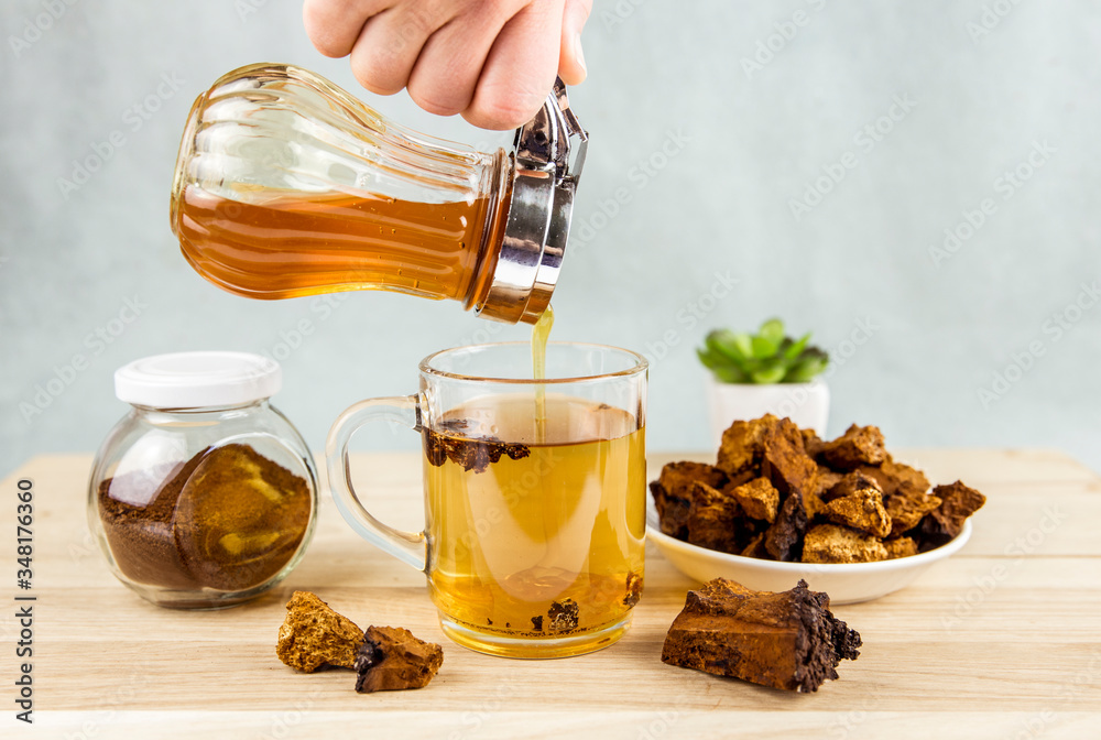 Person makind tea drink with wild natural Chaga mushroom from birch tree, Inonotus obliquus pieces in tea glass on wooden table indoors home. Alternative traditional medicine. Pouring honey in tea. Person makind tea drink with wild natural Chaga mushroom from birch tree, Inonotus obliquus pieces in tea glass on wooden table indoors home. Alternative traditional medicine. Pouring honey in tea.