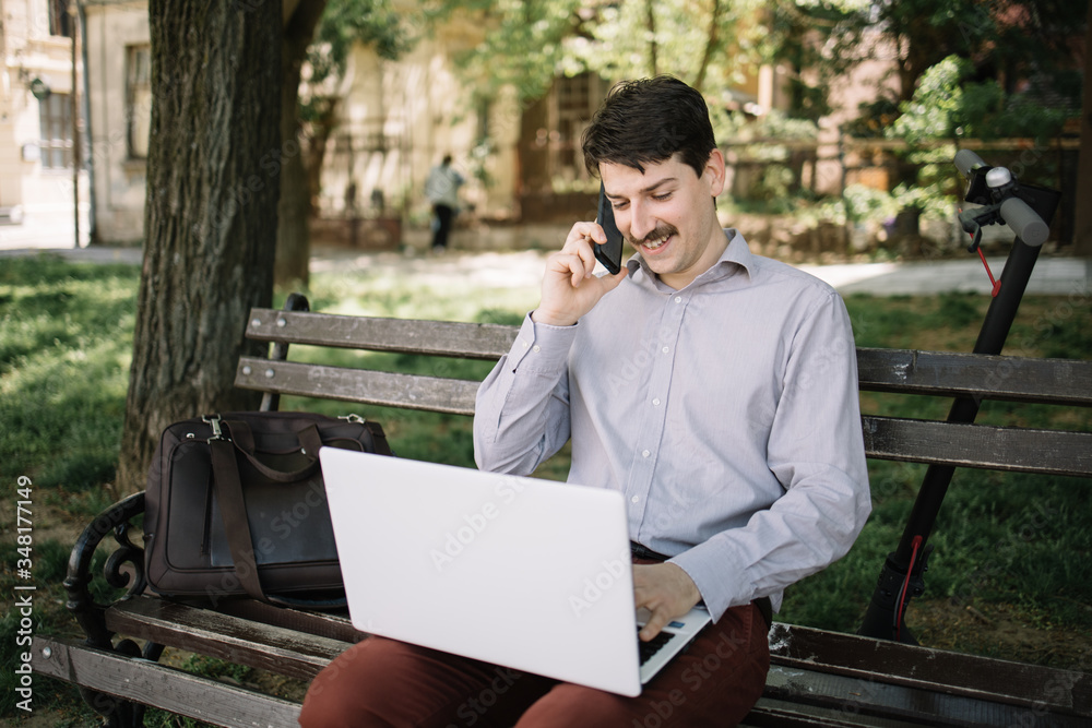 Smiling business man using laptop while talking on mobile. Young business man with moustache speaking on the phone while using laptop in park. Smiling business man using laptop while talking on mobile. Young business man with moustache speaking on the phone while using laptop in park.