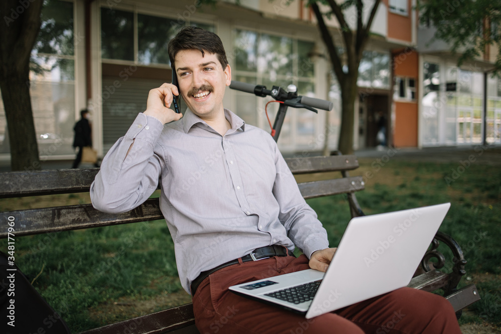 Guy holding phone while sitting on bench with laptop. Happy man with moustache sitting on bench with laptop and holding mobile phone outdoor. Guy holding phone while sitting on bench with laptop. Happy man with moustache sitting on bench with laptop and holding mobile phone outdoor.