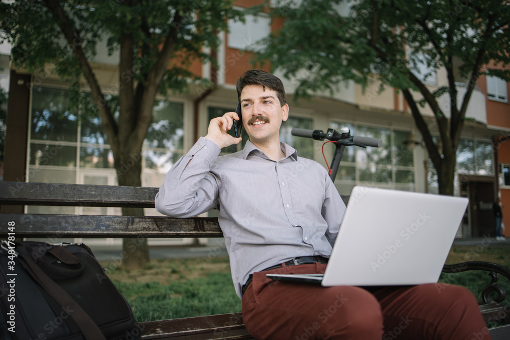 Man having conversation on the phone while resting on bench. Young man with moustache talking on the phone and sitting with computer on his lap. Man having conversation on the phone while resting on bench. Young man with moustache talking on the phone and sitting with computer on his lap.