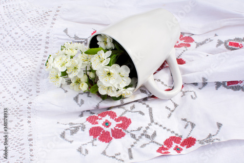 
A ceramic cup with flowers lies on a white tablecloth with embroidery. table setting with flowers. Rustic interior details.