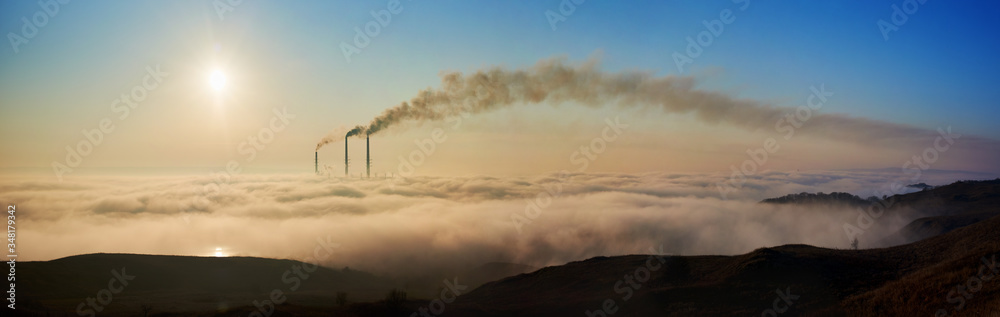 Panoramic view of a beautiful morning horizon with white clouds, rising sun and three pipes emissing dark smog into the atmosphere, concept of man-made disaster