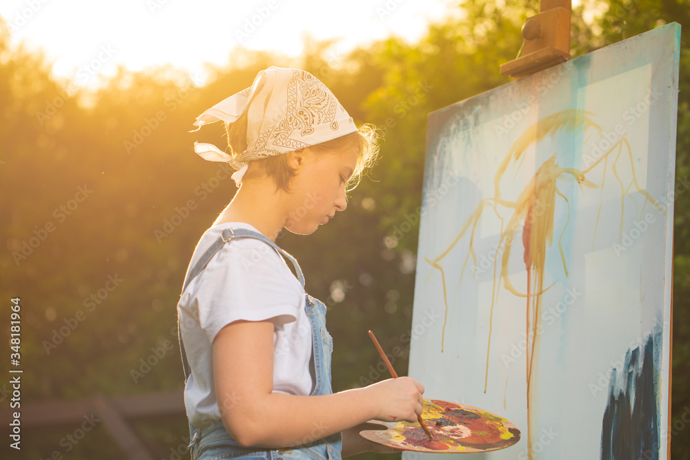 A young woman artist holds a brush and paints a picture on an easel in the rays of the sunset. The painter paints oil paintings in the garden