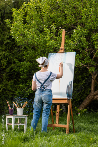 A young woman artist holds a brush and paints a picture on an easel in the rays of the sunset. The painter paints oil paintings in the garden