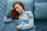 A red-haired girl in striped pajamas sleeps on a pillow, on blue bed linen. Frame on top