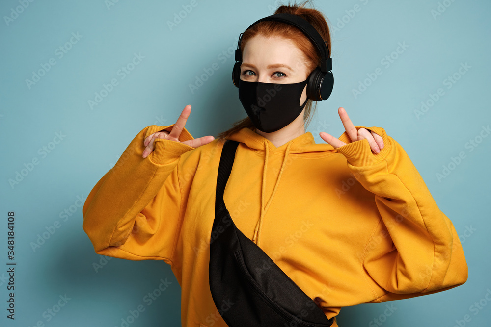 A red-haired girl in a yellow hoodie and a black mask stands on a blue background and shows her hands a peace gesture A red-haired girl in a yellow hoodie and a black mask stands on a blue background and shows her hands a peace gesture