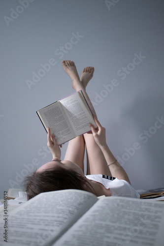 happy beautiful slim girl reading a book lying on the bed full of books, legs up. Grey bedroom and sheets. Homeschooling. Studying in quarantine. Reading is helpful- vertical photo