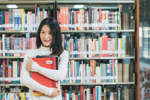 Asian young student in white casual suit reading the book in library of university or colleage.Standing and ้้้holding a book on a wooden table and bookcase in the background.Back to school