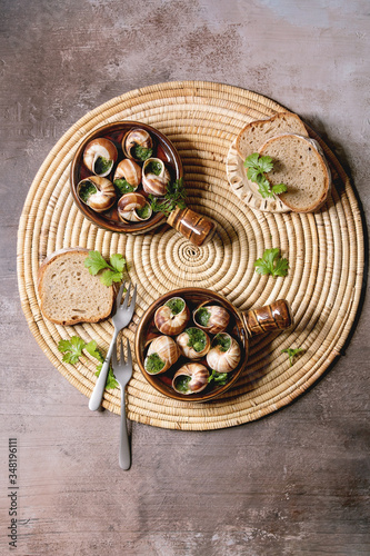 Escargots de Bourgogne - Snails with herbs butter, gourmet dish, in two traditional ceramic pans with parsley and bread on straw napkin over brown textured background. Flat lay, space