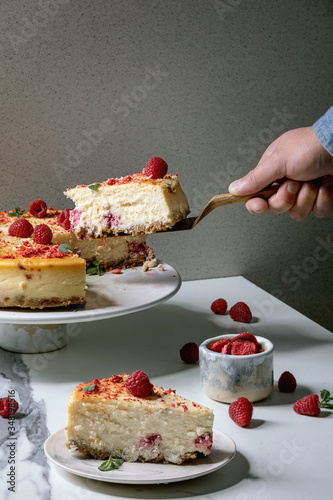 Man's hand hold piece of homemade raspberry baked cheesecake on wooden cake server. Portion on plate, fresh raspberries and mint above on white marble table photo