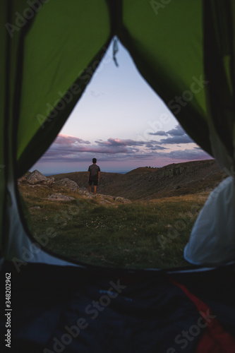 Man watching a sunset in the mountains viewed from inside a green tent.